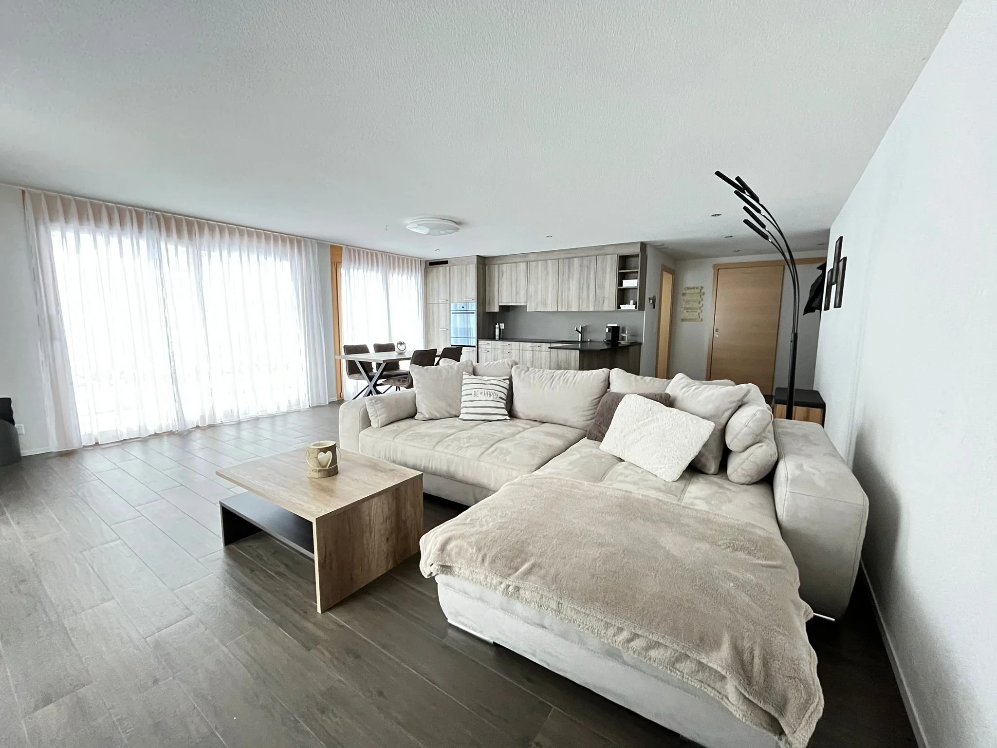 Offener Wohnraum mit einer großen beigen Couch, Couchtisch, Küchenbereich mit Holzschränken, Esstisch mit Stühlen, große Fenster mit weißen Vorhängen.