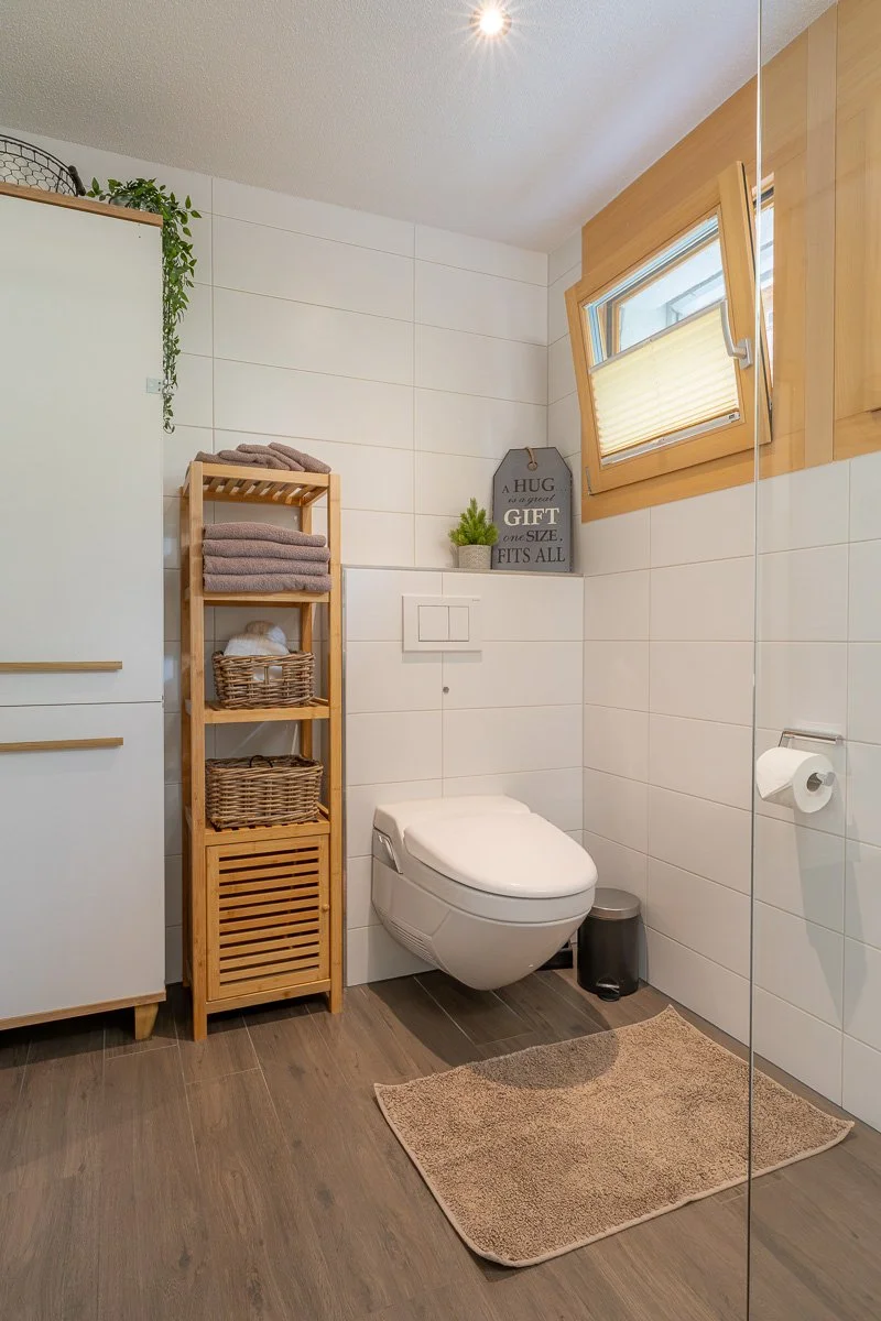 Modernes Badezimmer mit Toilettenschüssel, Holzregal, Handtüchern, Fenster und Duschabtrennung.