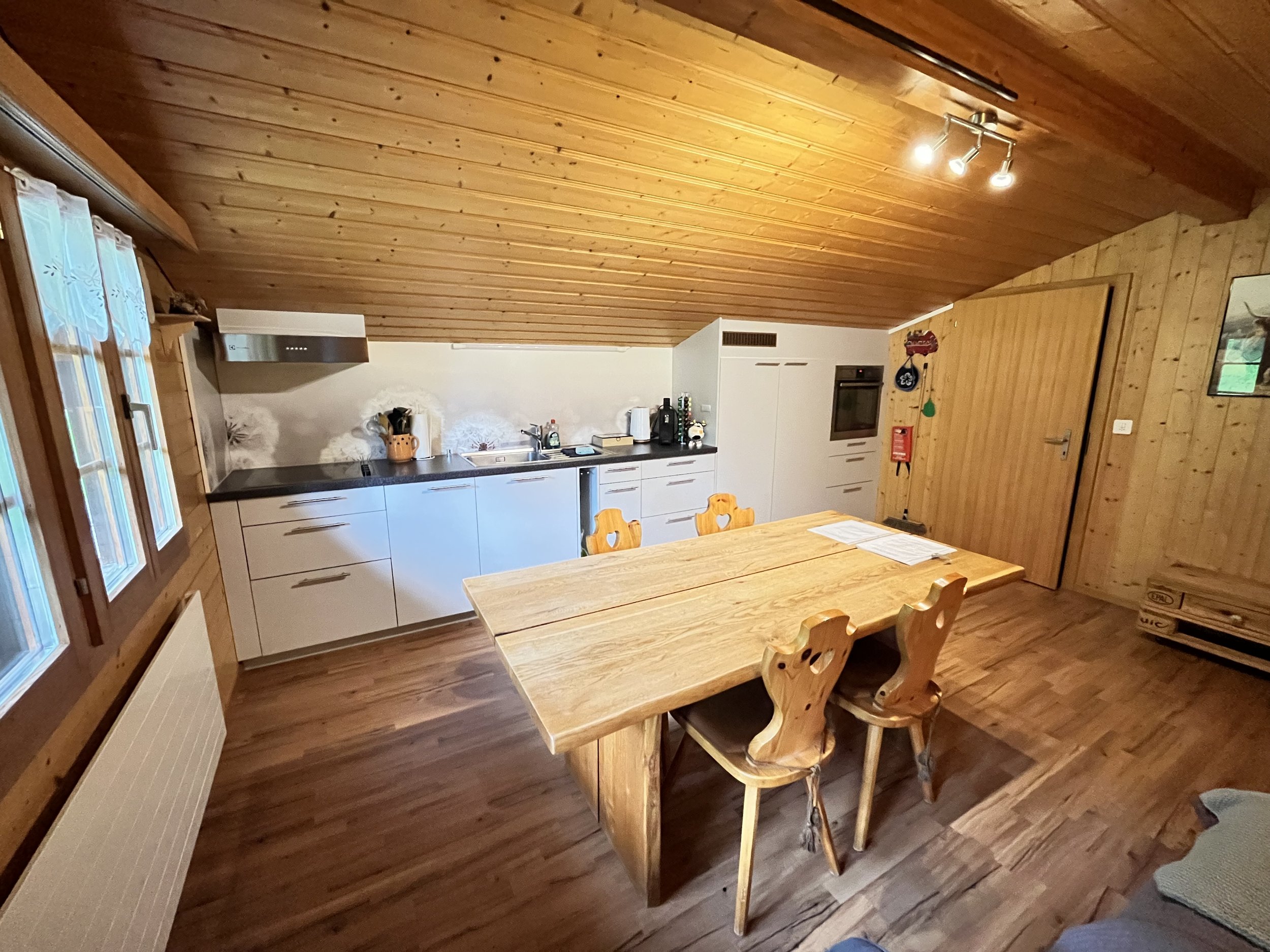 Ein rustikaler Küchenraum mit Holztischen, weißen Schränken und einem Holzpaneel-Deckenbereich, große Fenstern mit Gardinen, und Küchengeräten auf der Arbeitsplatte.