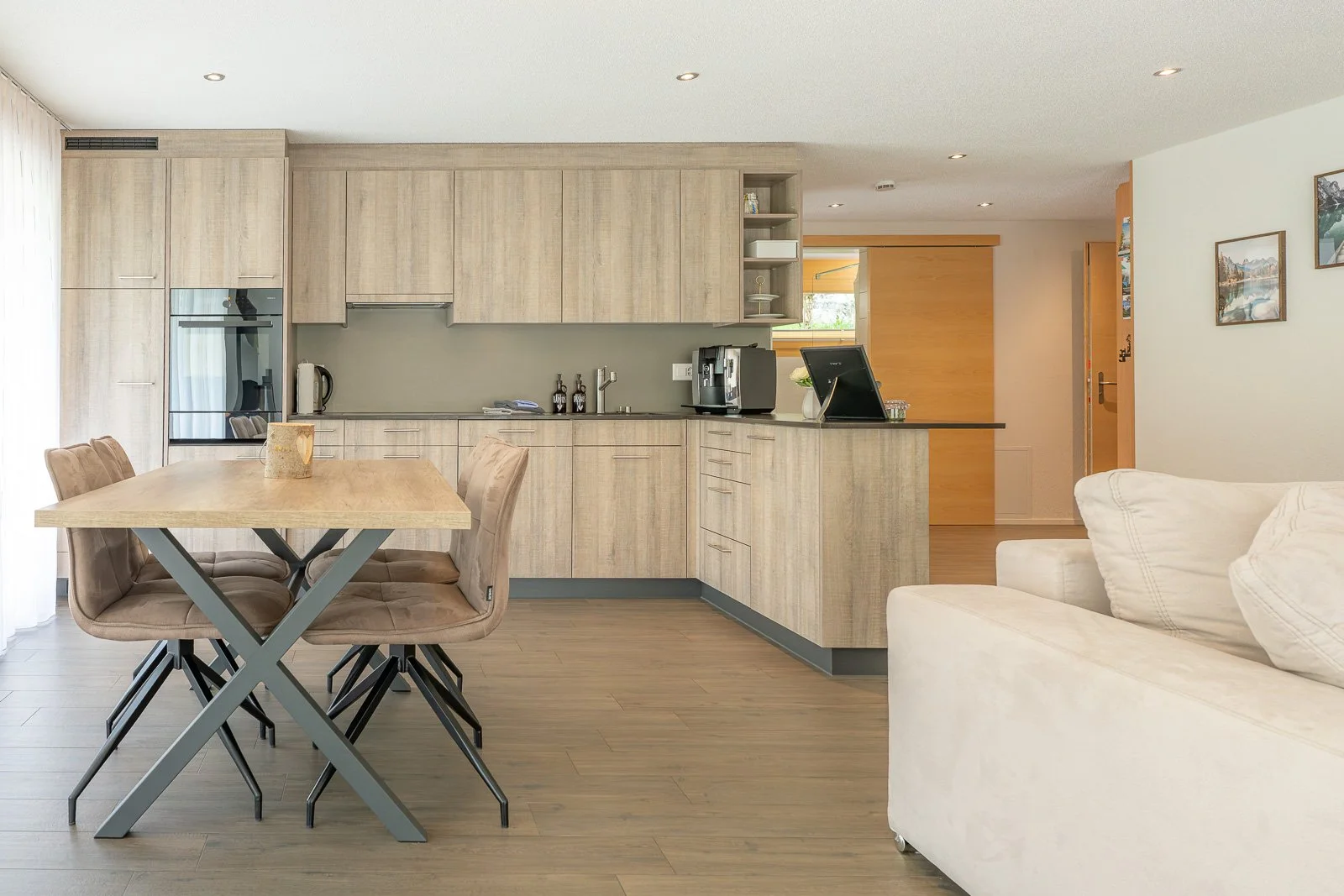 Offene Küche mit Esstisch und Stühlen, weißes Sofa, Wände mit Bildern, Holzfußboden, helle Einrichtung.