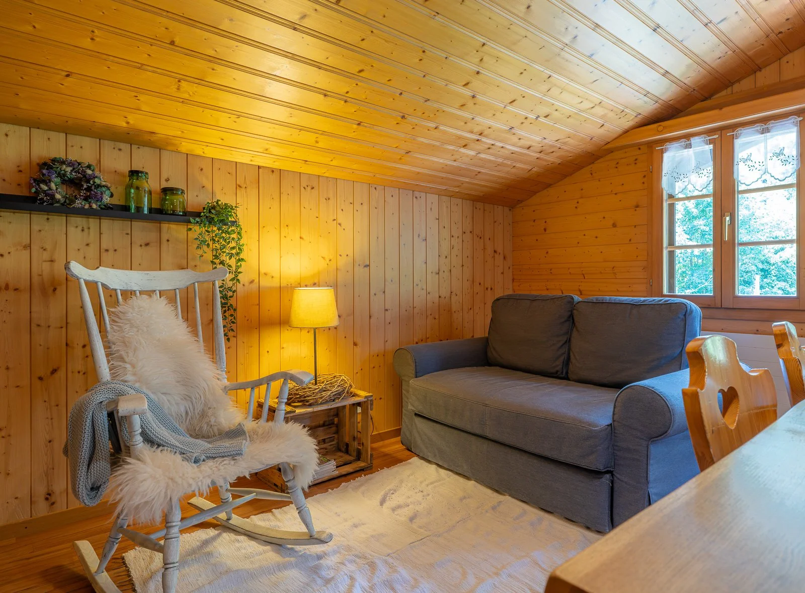 Gemütliches Wohnzimmer mit Holzverkleidung, grauem Sofa, Schaukelstuhl mit Fellüberwurf, Stehlampe, Fenster mit Vorhängen und Deko-Accessoires auf einem Regal.