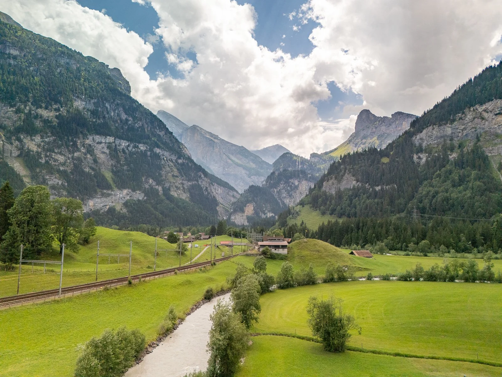 Grünes Tal in den Alpen mit Fluss, Häusern, Wiesen und Bergen im Hintergrund, teilweise bewölkter Himmel.