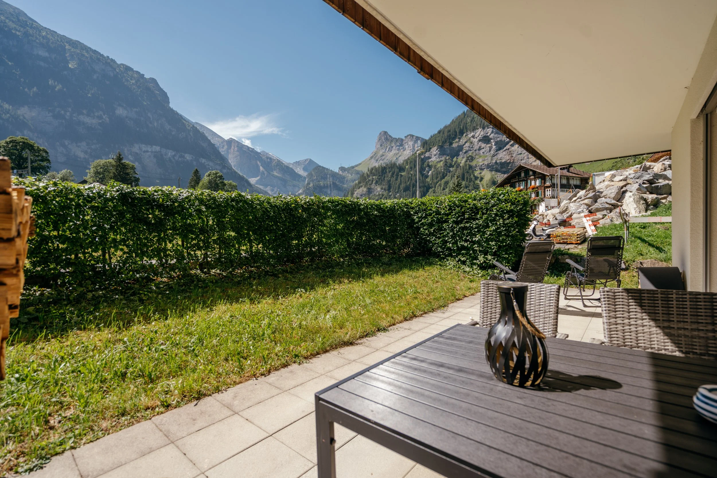 Balkon mit Gartenmöbeln, Blick auf Berge und Himmel, Büschen, Rasen, und ein Haus im Hintergrund in einer alpinen Gegend.
