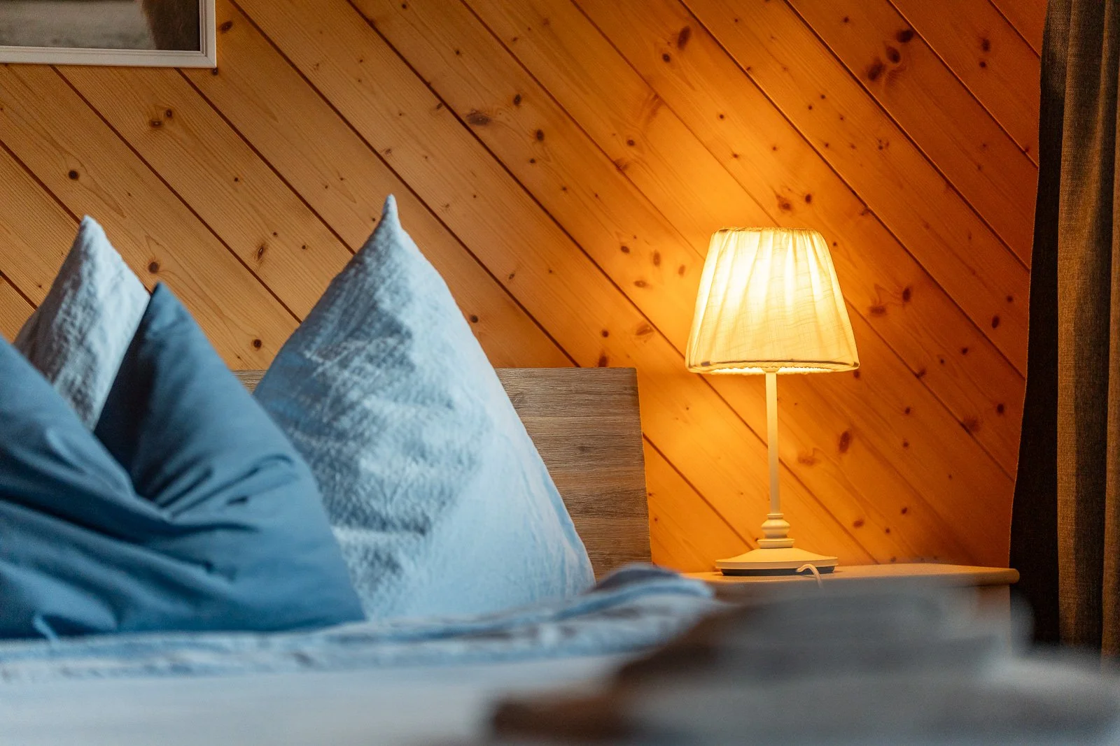 Bett mit blauen Kissen und einer Nachttischlampe, helle Holzvertäfelung an der Wand im Schlafzimmer.