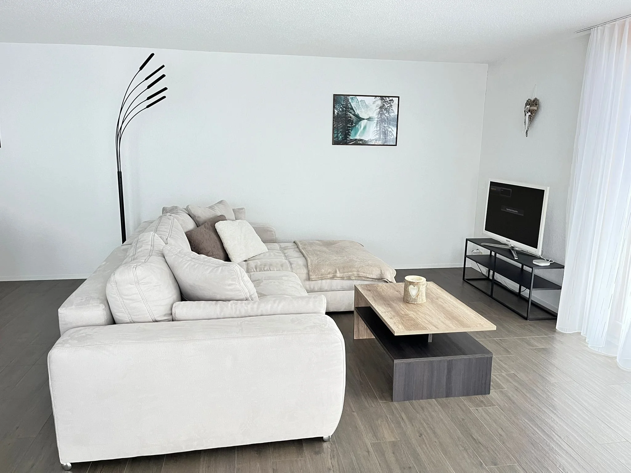 Moderner, heller Wohnzimmerbereich mit einer weißen Couch, einem schwarzen Beistelltisch, einem Fernseher auf einem Gestell, Wanddekorationen und einem Bild an der Wand, Laminatboden und Vorhängen auf der rechten Seite.
