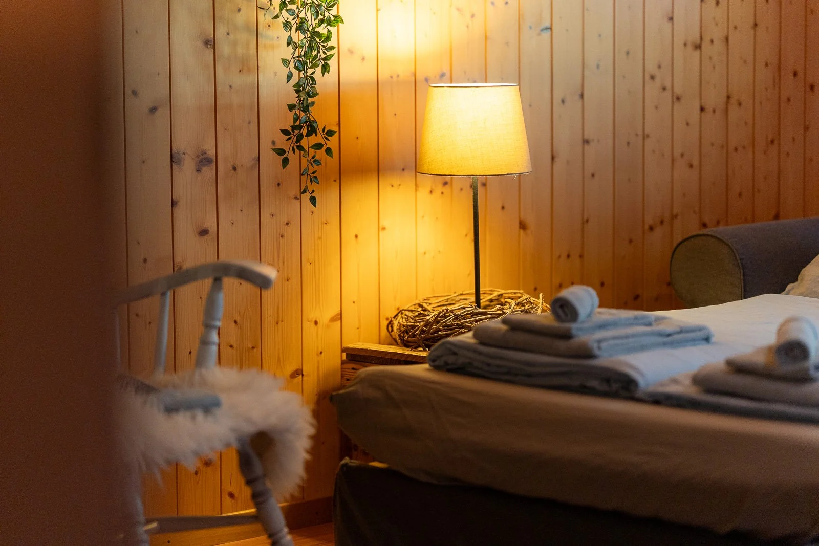 Bett im rustikalen Holzhaus, mit ordentlich gefalteten Handtüchern auf der Bettdecke, seitlich eine Stehlampe mit warmem Licht, Holzverkleidung an der Wand, Pflanzen im Hintergrund.