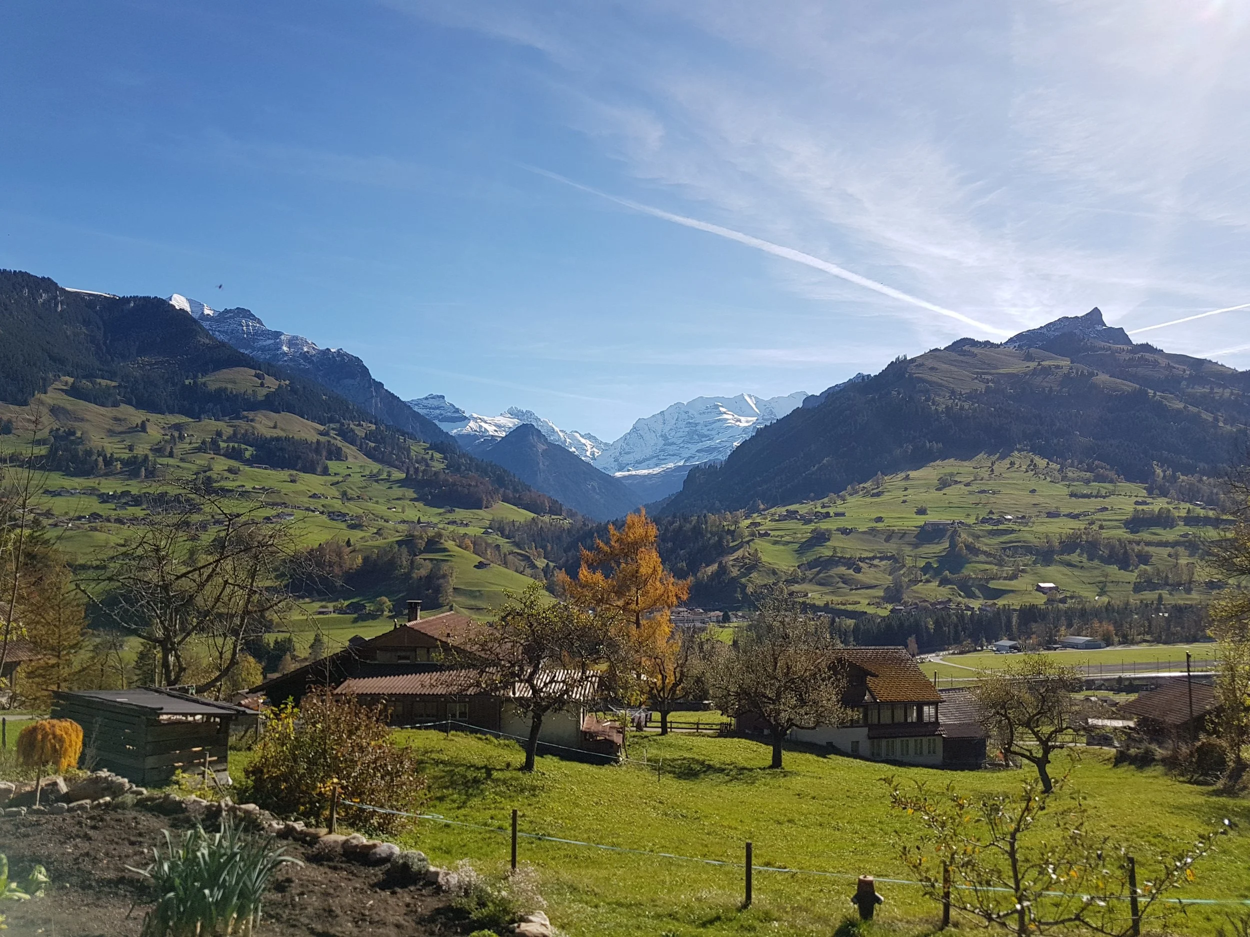 Berglandschaft mit grünen Hügeln, Häusern und schneebedeckten Gipfeln im Hintergrund bei sonnigem Wetter.