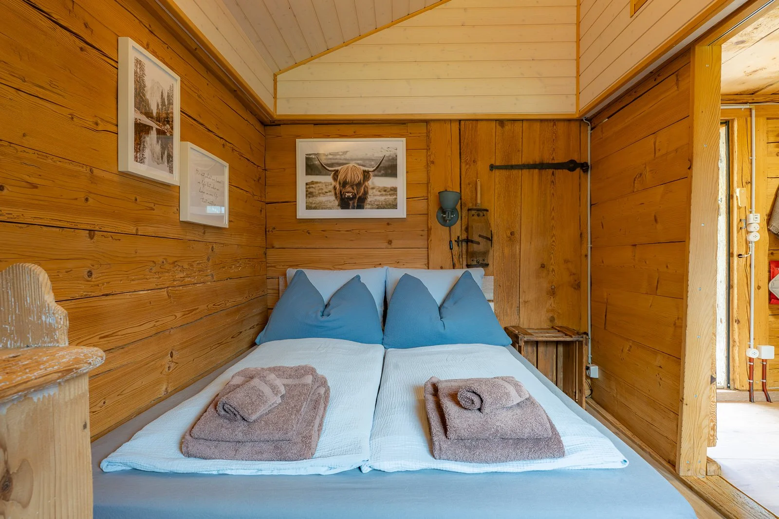 Ein Schlafzimmer mit Holzverkleidung, einem Bett mit blauen Kissen, auf dem zwei Handtücher liegen, und Bildern an der Wand, darunter ein Bild eines Hochlandrinds.