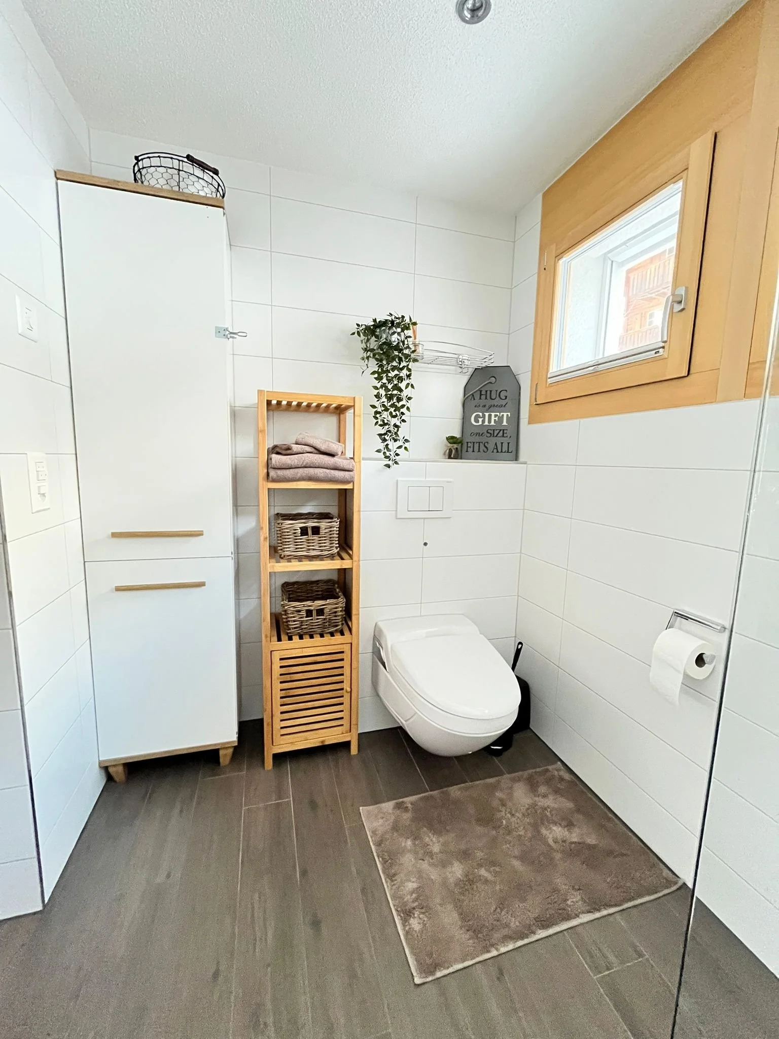 Grab der Badezimmer mit weißen Fliesen, einem Fenster mit Holzrahmen, einem weißen WC, einem weißen Schrank, einem Holzregal mit Handtüchern und Körben, einer Pflanze, einem Wanddekor und einem Toilettenpapierhalter.