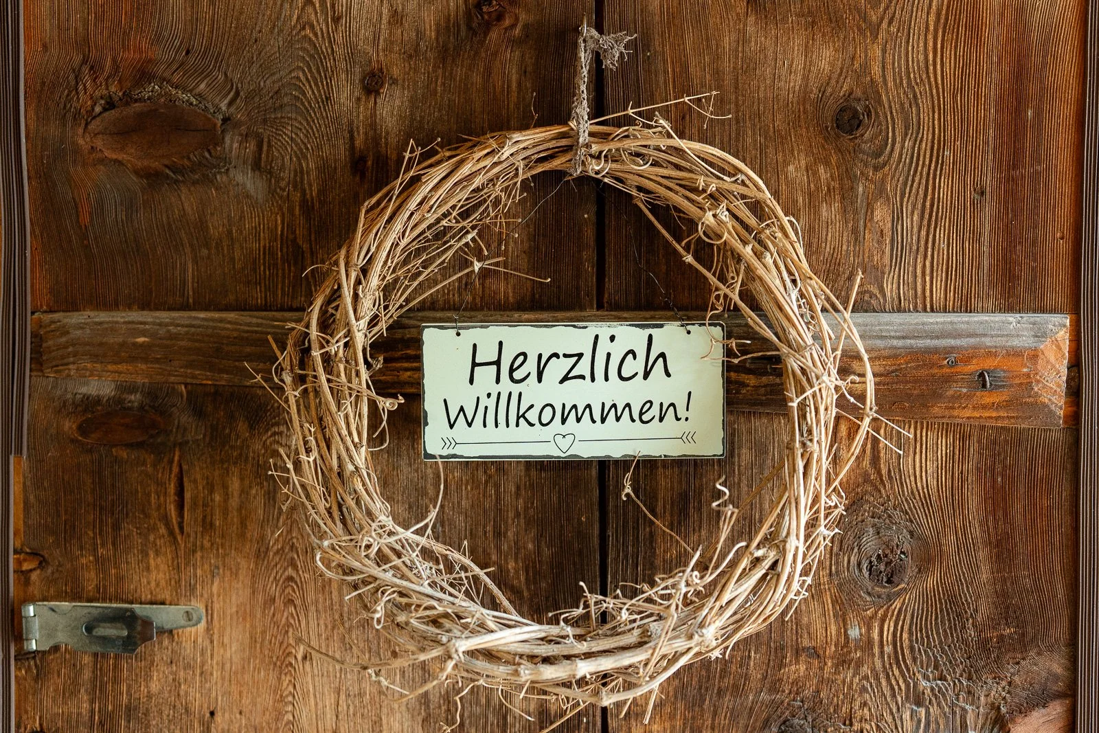 Ein Holzschild mit der Aufschrift 'Herzlich Willkommen!' hängt in einem Kranz aus Zweigen an einer Holztür.