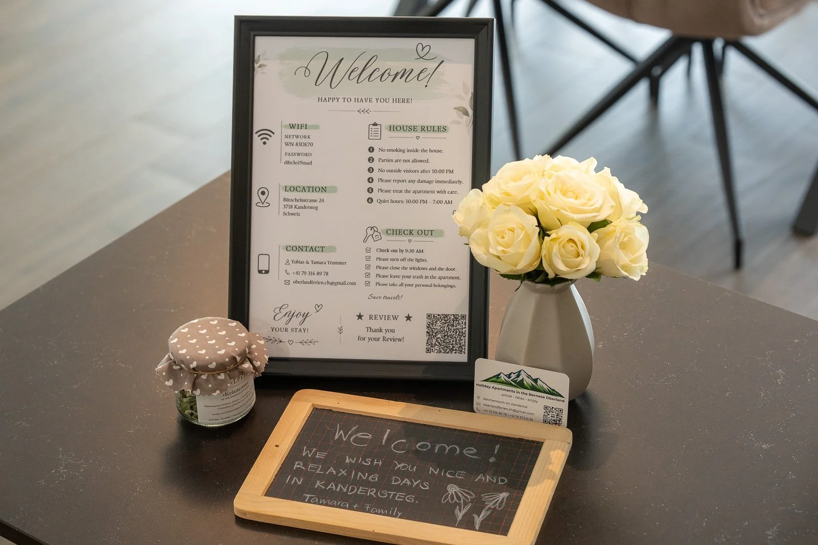 Ein Willkommensschild mit WLAN- und Hausregeln, einem Blumenstrauß in einer Vase, einer kleinen Flasche mit Kräutern, einer Tafel mit Willkommensnachricht, und einer Visitenkarte auf einem grauen Tisch.