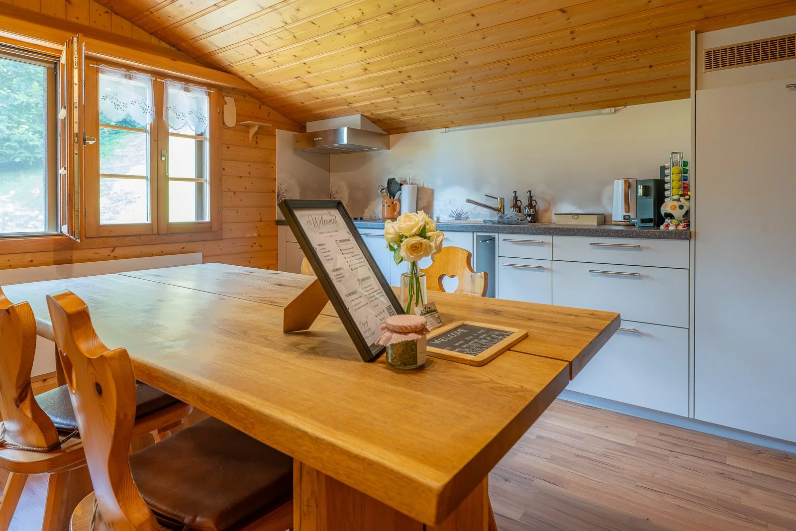Gemütliche Küche mit Holzdecke und Fenster, Holztisch mit Blumen, Menükarten und Gewürzglas, weiße Schränke, Küchengeräte und Zubehör.