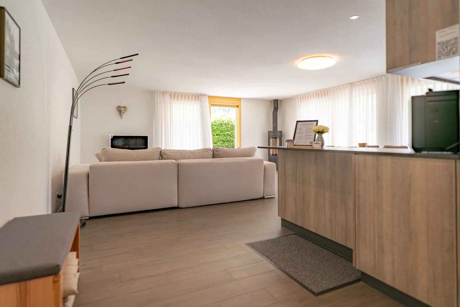 Ein gemütliches Wohnzimmer mit einem großen weißen Sofa, Vorhängen vor den Fenstern, einem Kamin, einem Ecktischlampe, einem kleinen Fernseher und einer Küchenbar mit Blumen auf der Theke.