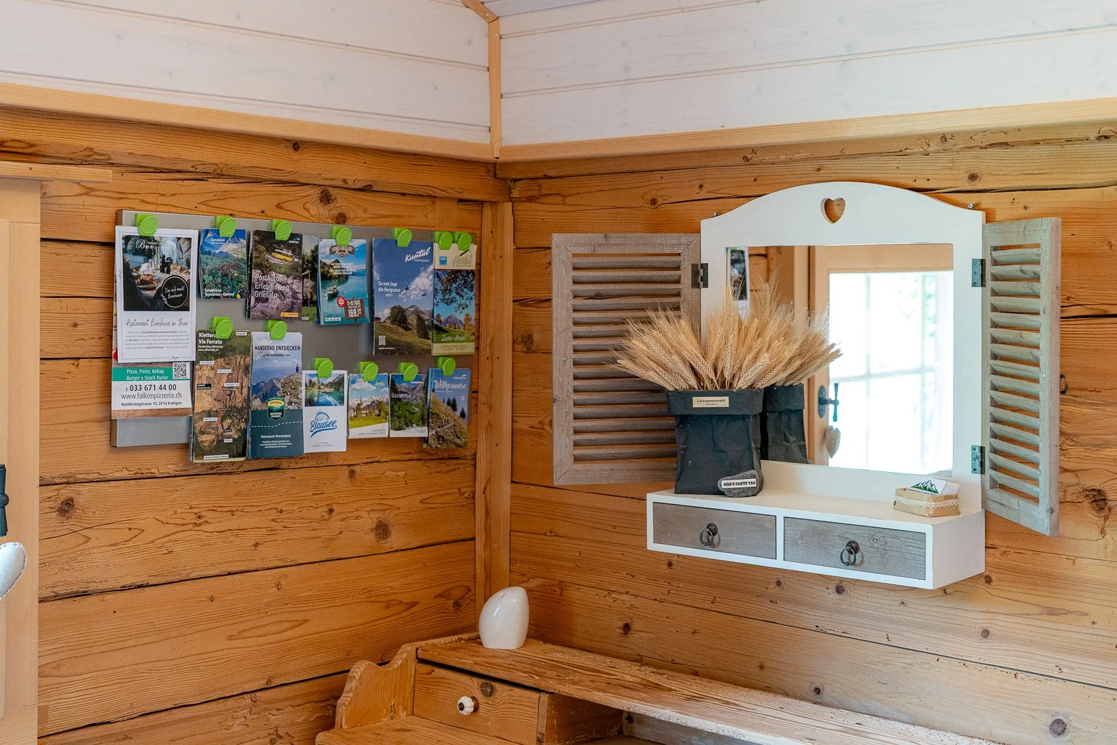 Holzverkleideter Raum mit an einer Wand angebrachter Postkarten- und Informationsflächen, einem Wandspiegel mit geöffneten Shutters, Dekoration mit Trockenblumen in einer schwarzen Vase und einem kleinen Scallop-Boxen.