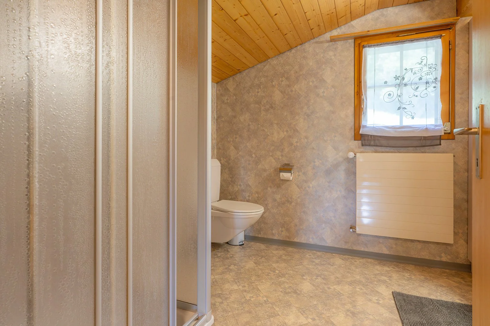 Badezimmer mit Toilette, Heizkörper und Fenster mit Gardine, Holzdecke, Fliesenboden und Duschkabine