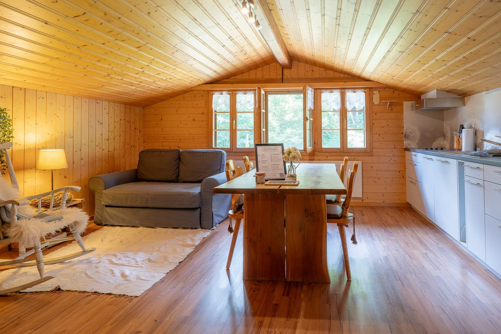 Ein gemütliches Wohnzimmer in einer Holzhütte mit einem grauen Sofa, einem Kinderrocker, einem Esstisch mit Stühlen, einer Vase mit Rosen, drei Fenster, einer kleinen Küche mit weißen Schränken und einem Herd, Holzboden und Holzwänden.