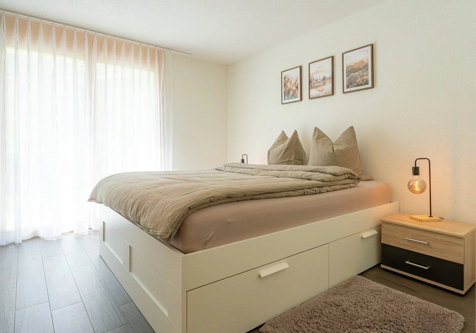 Ein gepflegtes Schlafzimmer mit einem großen Bett, beige Bettwäsche und Kissen, kleines Nachttisch mit einer modernen Lampe, drei Wandbilder über dem Bett, große Fenster mit weißen Gardinen, helles Zimmer