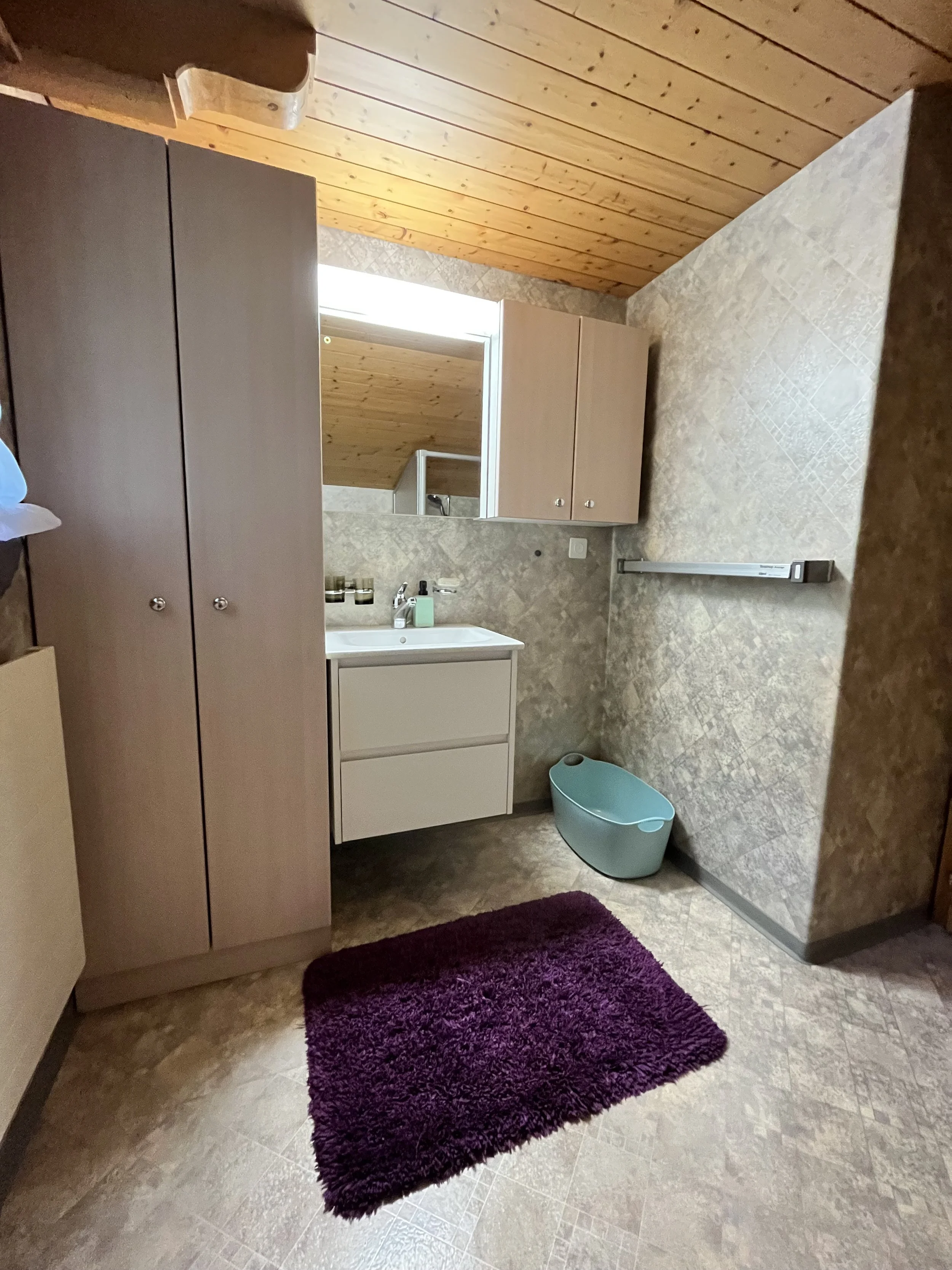 Badezimmer mit Holzdecke, grauen Wänden und Teppich. Es gibt einen Spiegel, eine Waschbeckenunterschrank, eine Dusche, einen Badeeimer und einen dunkelvioletten Teppich.