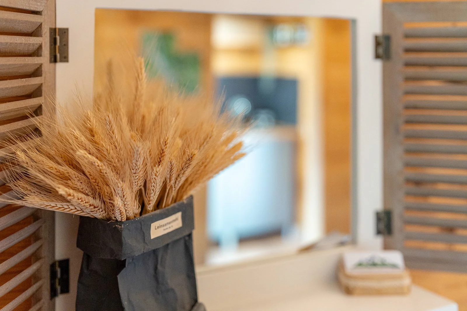 Trockenblumen in einer schwarzen Vase vor einem Spiegel, im Hintergrund eine offene Holzklappluke.
