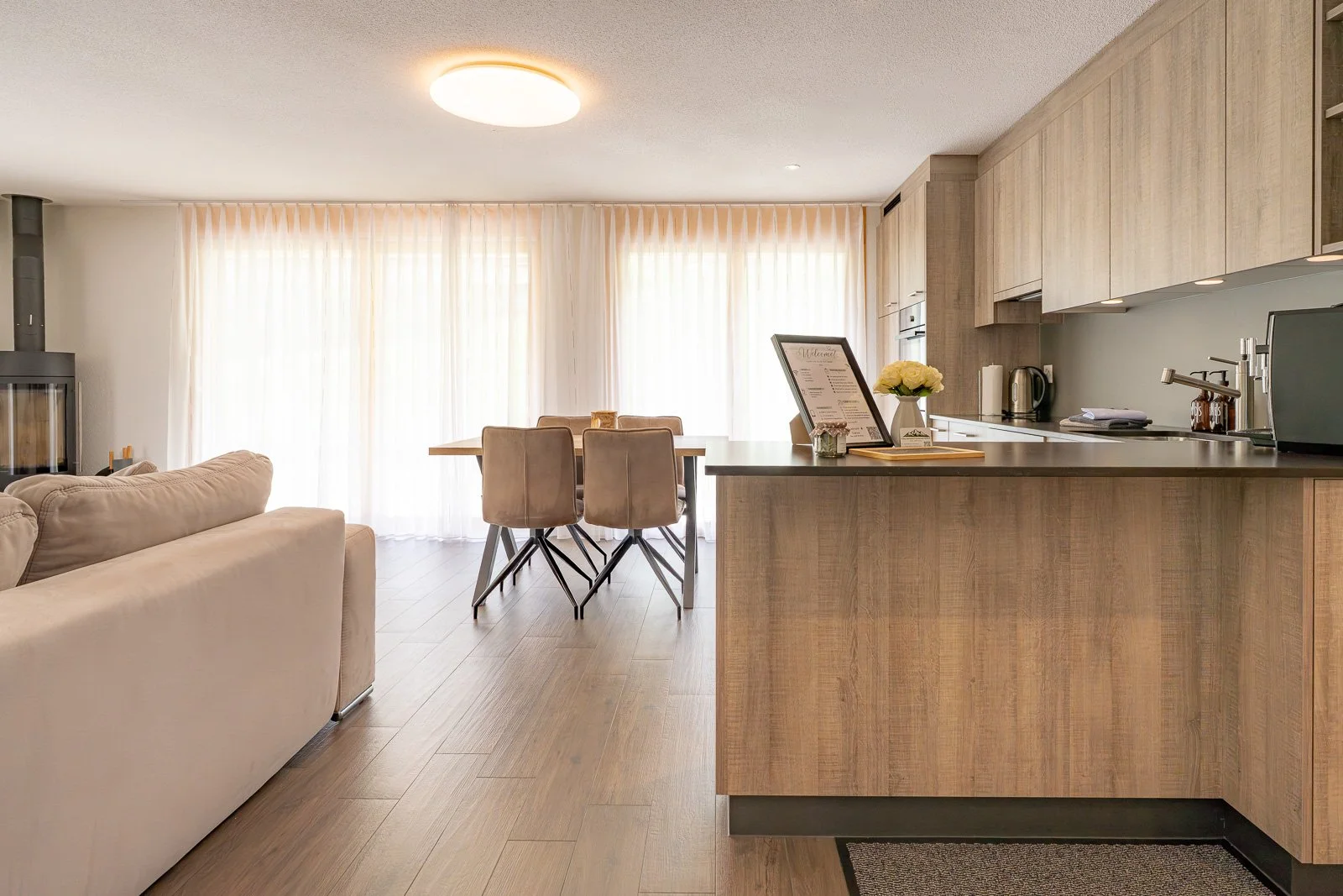 Modernes, helles Wohnzimmer mit offener Küche, Blick auf Esstisch mit vier Stühlen, Couch, große Fenster mit weißen Vorhängen, Holzfußboden, minimalistische Einrichtung.