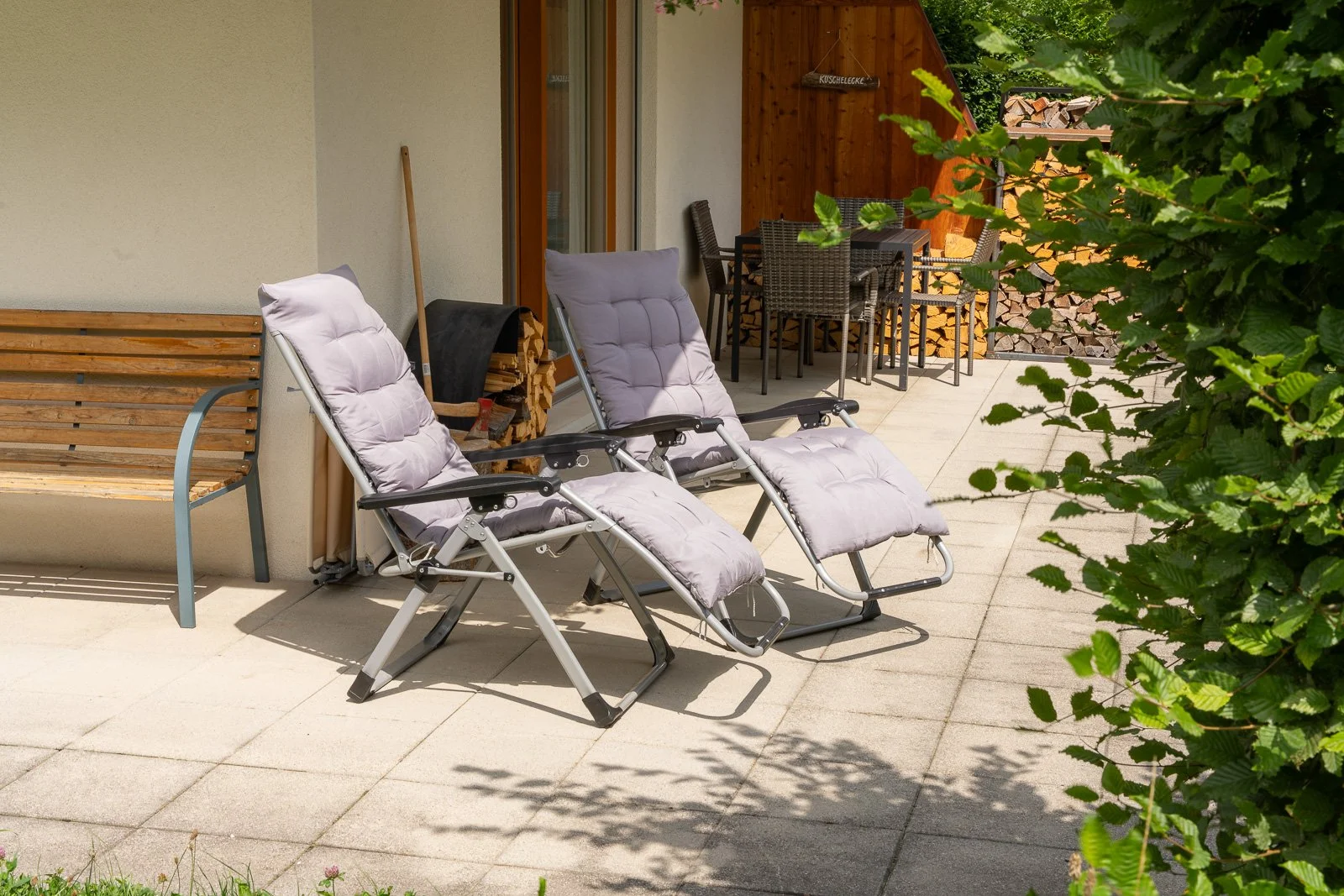 Ein Terrassensofa mit zwei Liegestühlen, einen Holzbank, einem Kamin und einem Essbereich mit einem Tisch und Stühlen, umgeben von grünen Bäumen.