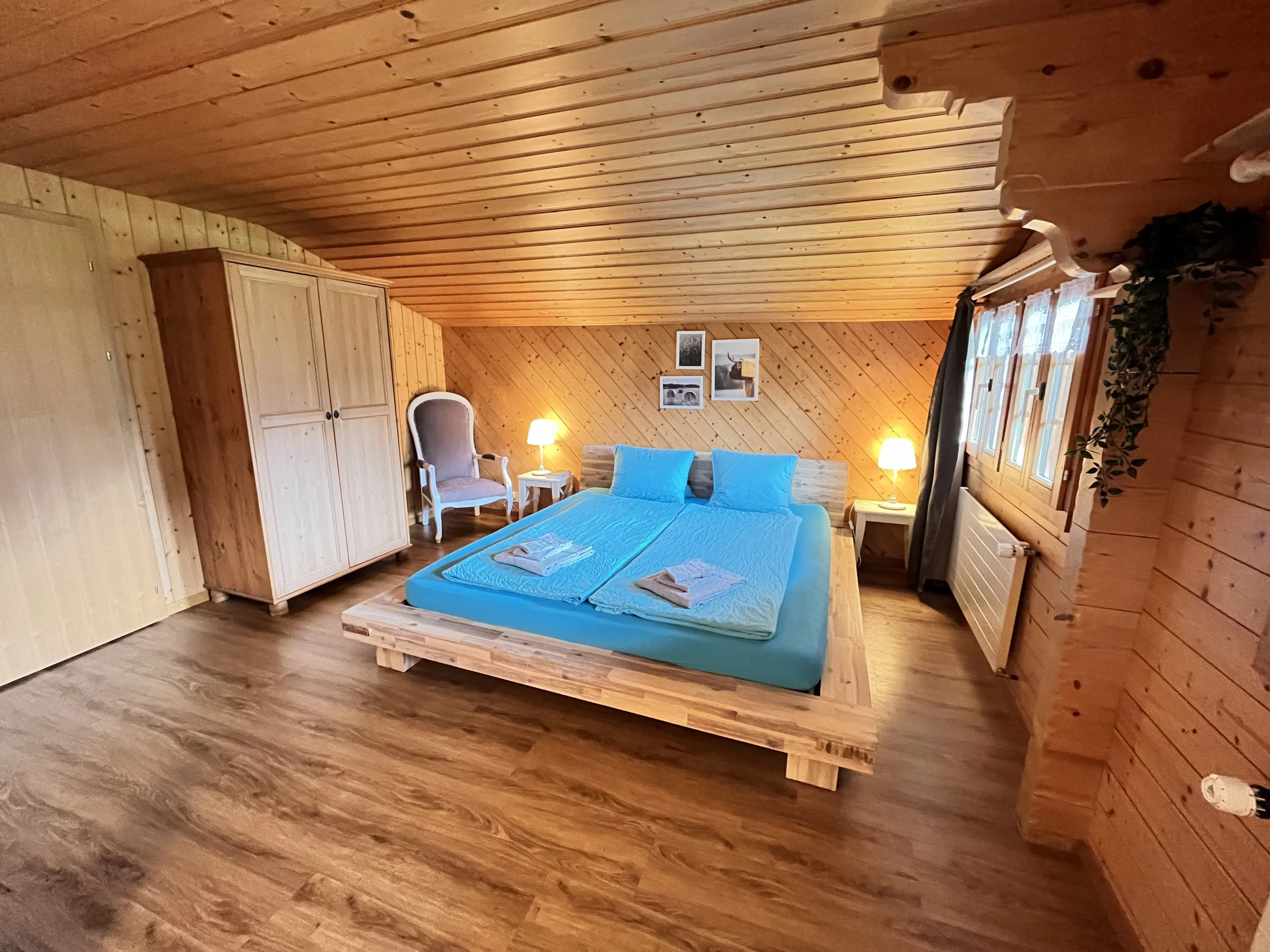 Ein gemütliches Schlafzimmer mit Holzvertäfelung, einem Doppelbett mit blauer Bettwäsche, zwei Nachttischen mit Lampen, einem hohen Kleiderschrank, einem акхел (Sessel) und Bildern an der Wand, natürlichem Licht durch Fenster.
