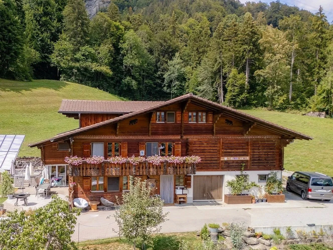 Ein Holzhaus mit Balkon, blühenden Blumen und einem Garten, umgeben von grünen Hügeln und einem bewaldeten Hügel im Hintergrund.
