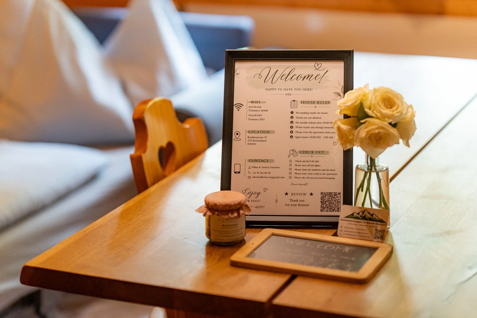 Ein Tisch mit einem Blumentopf mit weißen Rosen, einer Tafel mit Informationen, einem kleinen Joghurtglas, einer kleinen Tafel mit der Aufschrift "Welcome" und einem QR-Code, in einer gemütlichen, schön dekorierten Umgebung.