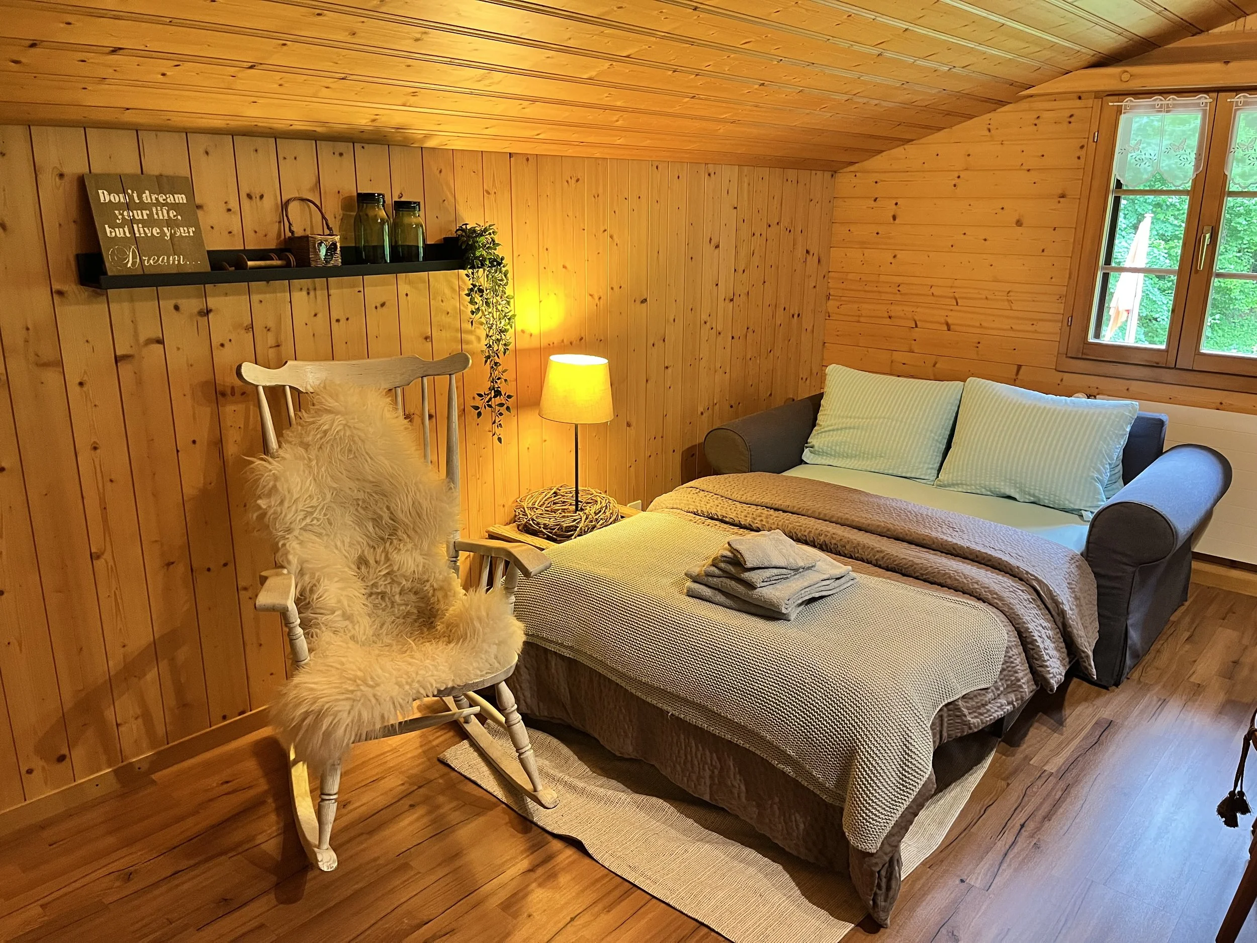 Gemütliches Schlafzimmer mit Holzpaneelwänden, einem Bett, einem Sessel mit Fell, einem Nachttisch mit Lampe und einem Fenster nach draußen.