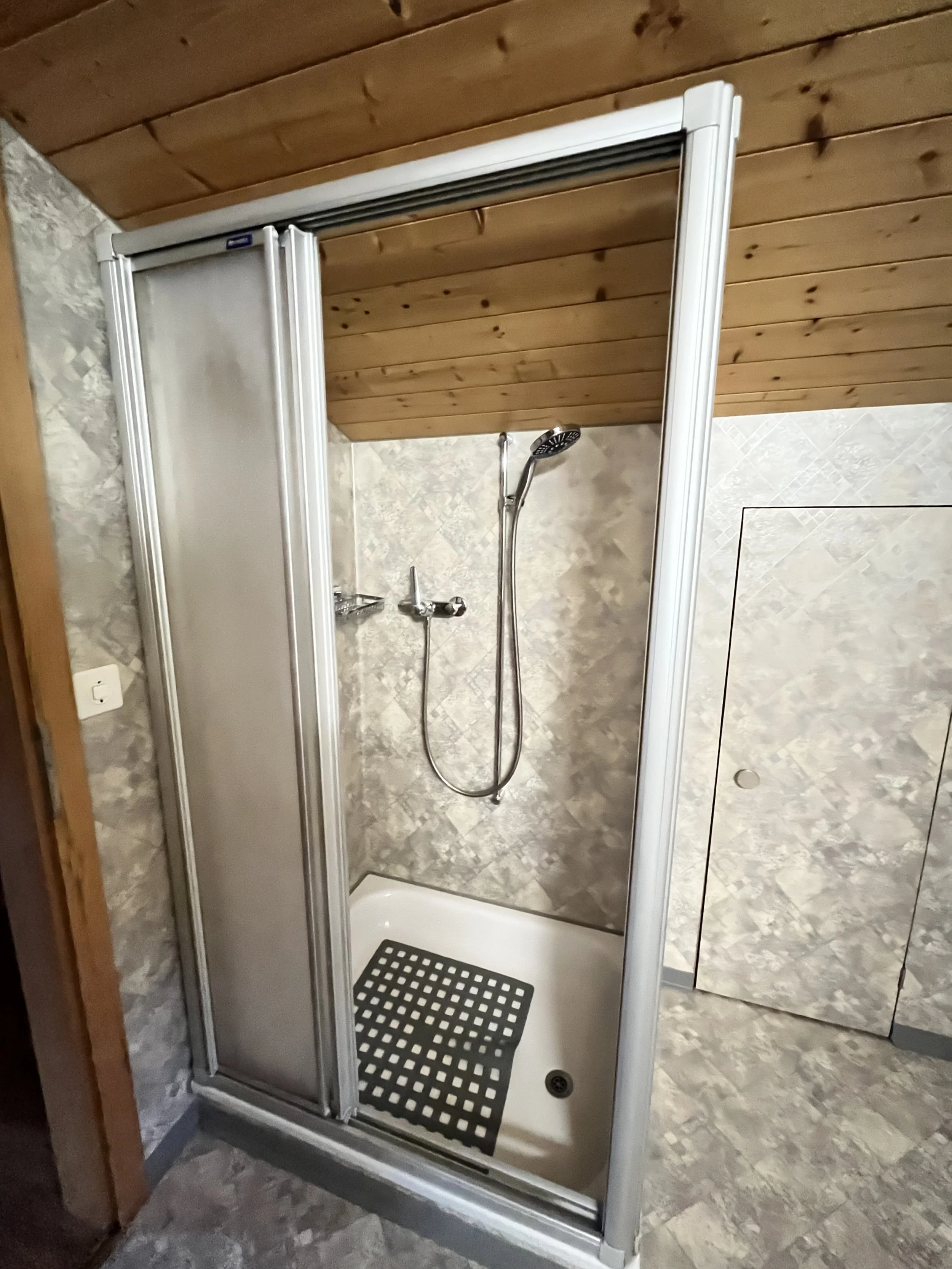 Kleines Duschkabinen mit einem schwarzen rutschfesten Mattenabsatz, ein Handbrausekopf an der Wand, Holzdecke und Fliesen auf Wänden und Boden.