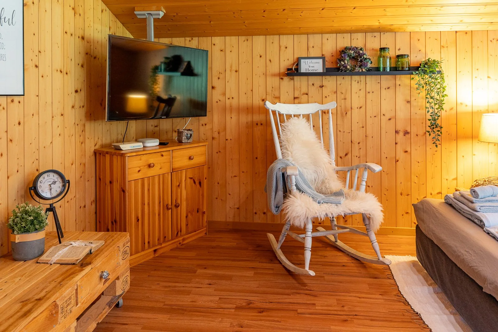 Ein Wohnzimmer mit Holzvertäfelung, einem Fernseher auf einem Holzmöbel, einem weißen Schaukelstuhl mit Kissen, einem Bett mit gestapelten Handtüchern, einem kleinen Tisch mit Uhr, Buch und Pflanze, und einer Wandregal mit Dekorationen.