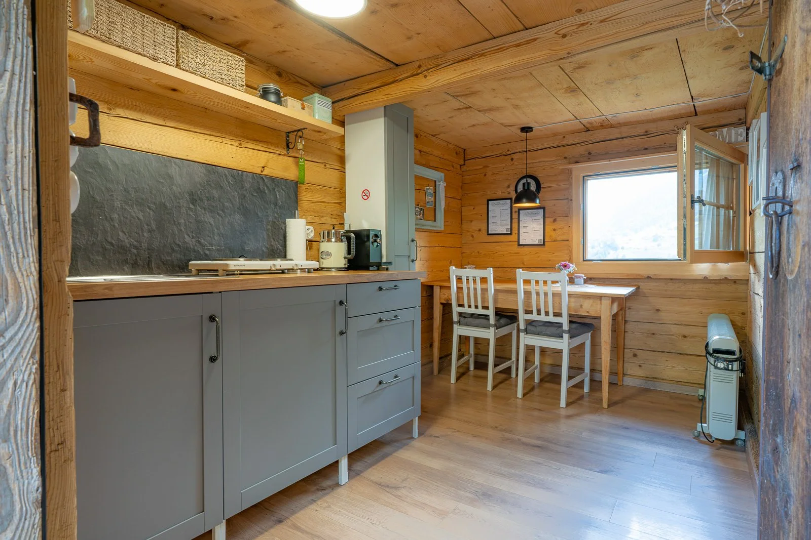 Gemütliche Holz-Küche mit grauen Schränken, Küchenutensilien, Fenster und Esstisch mit drei weißen Stühlen in einem Blockhaus.