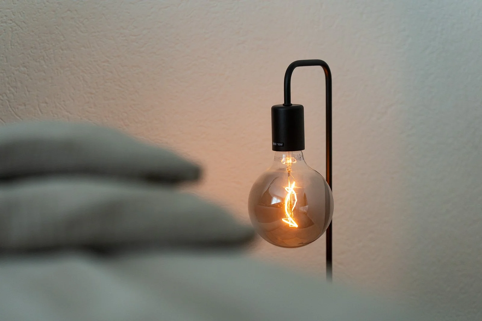 Ein beleuchtete Glühbirne mit sichtbar dimmmer Filament, hängt an einem schwarzen Lampenarm vor einer hellen Wand.
