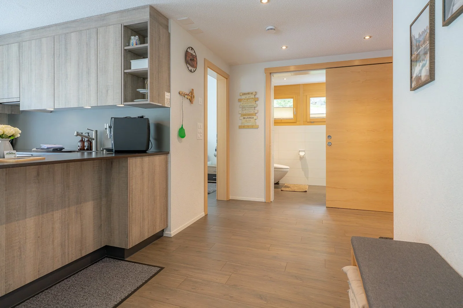 Innenraum eines Hauses mit Küche, einem Badezimmer und einem Eingangsflur, Holzboden, helle Wände, Fenster und Dekorationen.