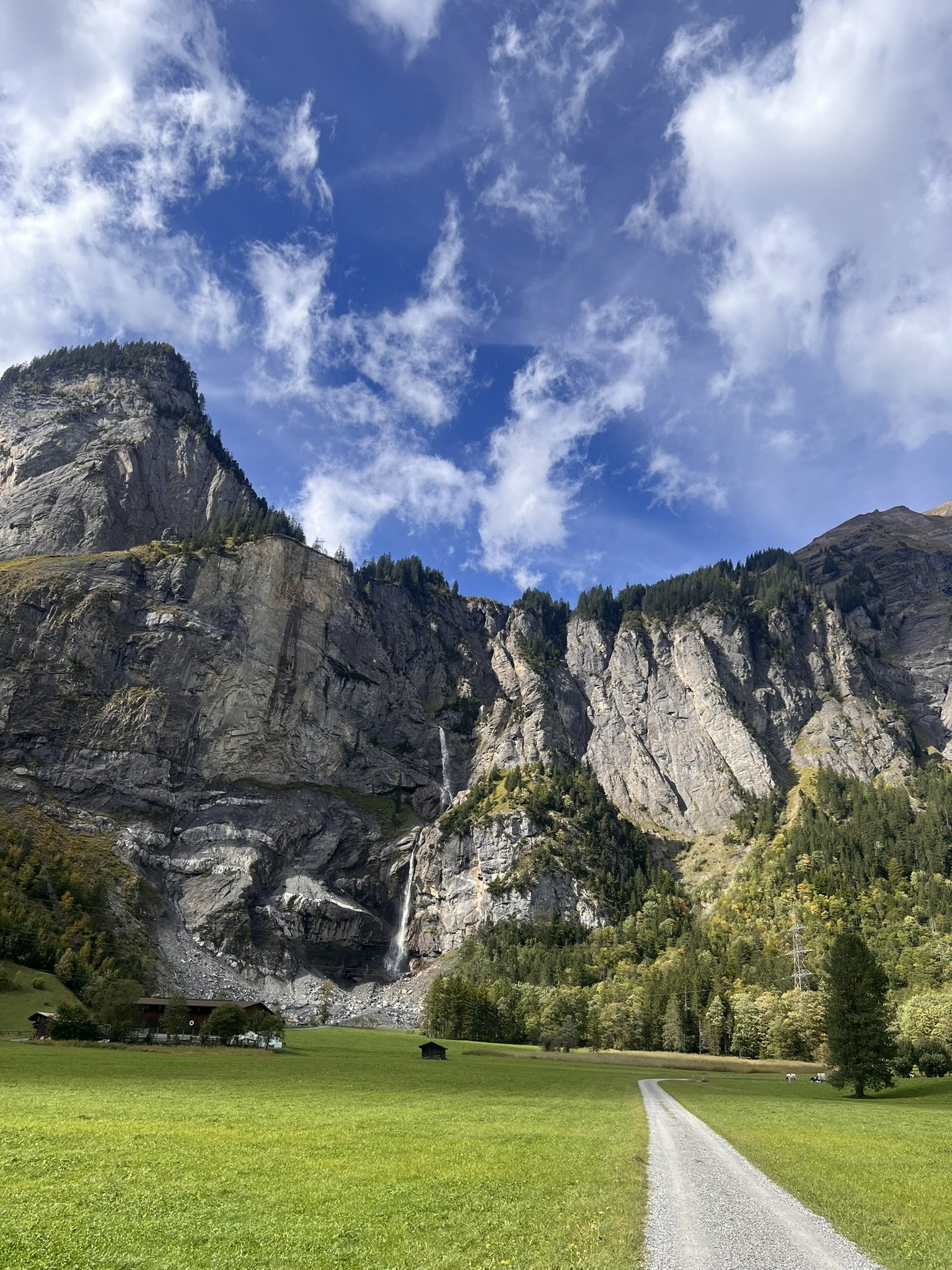 Berglandschaft mit Wasserfall, grüne Wiesen und Wald, blauer Himmel mit Wolken.