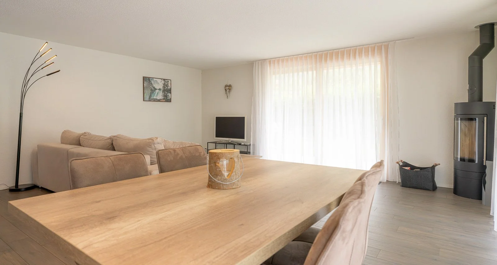 Moderner, heller Wohnzimmer mit einem großen Holztisch, bequemen Sofas, Kamin und Fernseher, große Fenster mit weißen Vorhängen.