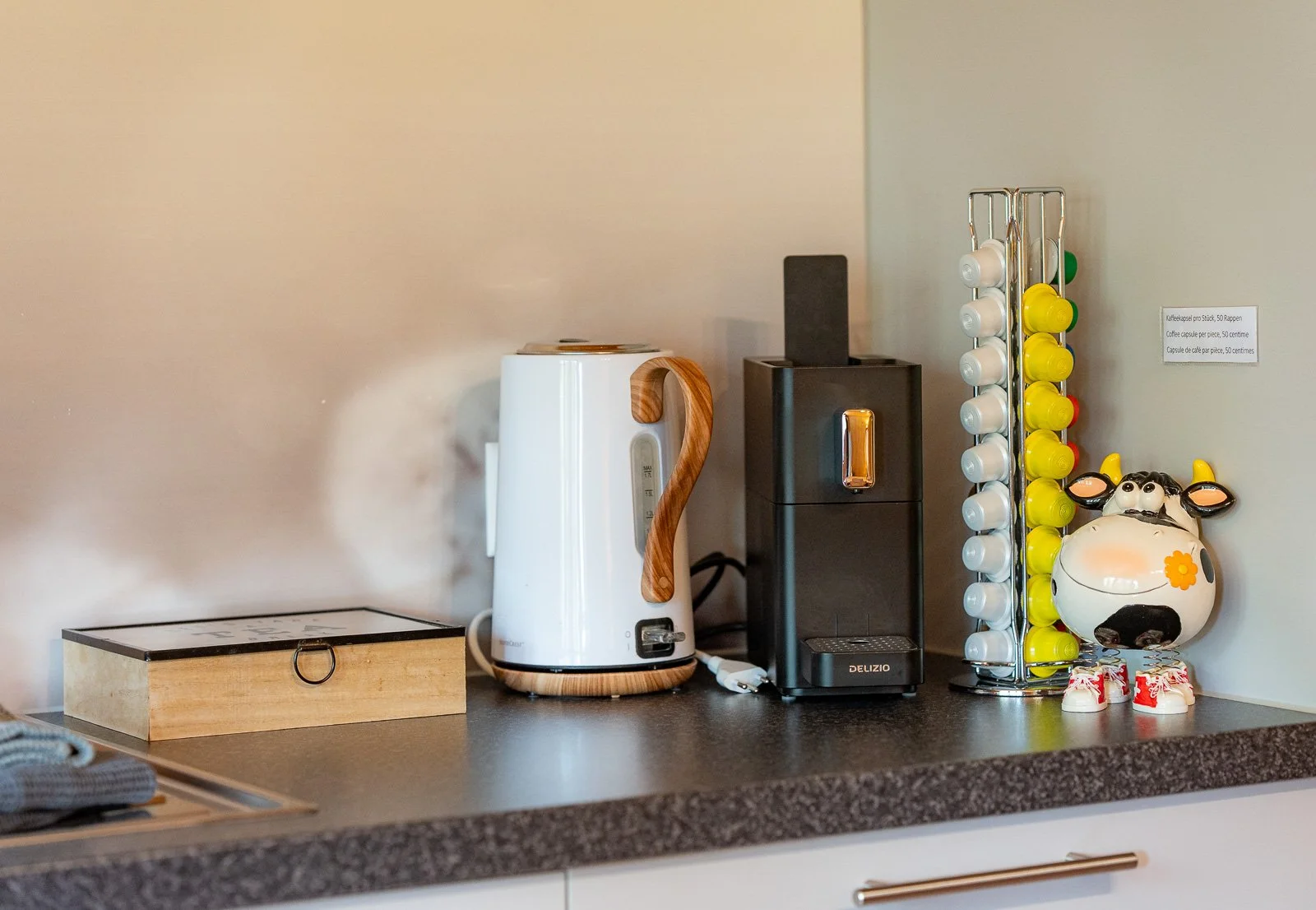 Supermarktküche mit Wasserkocher, Kaffeemaschine, Kapselspender, Cow-Decor, und kleinem Holzkästchen auf der Arbeitsfläche.