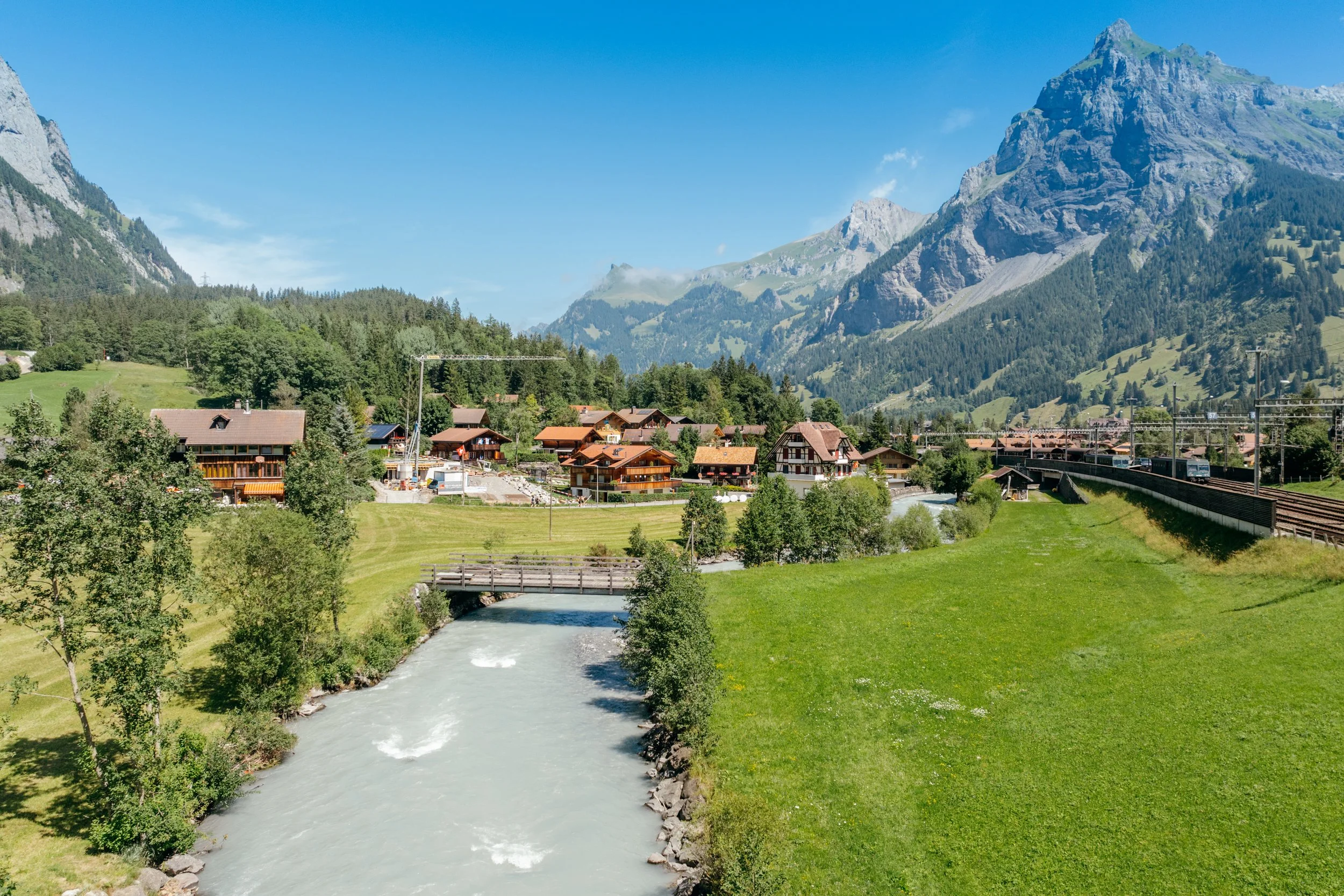 Eine malerische alpine Landschaft mit einem Fluss, grünen Wiesen, traditionellen Holzhäusern und hohen Bergen im Hintergrund bei klarem Himmel.