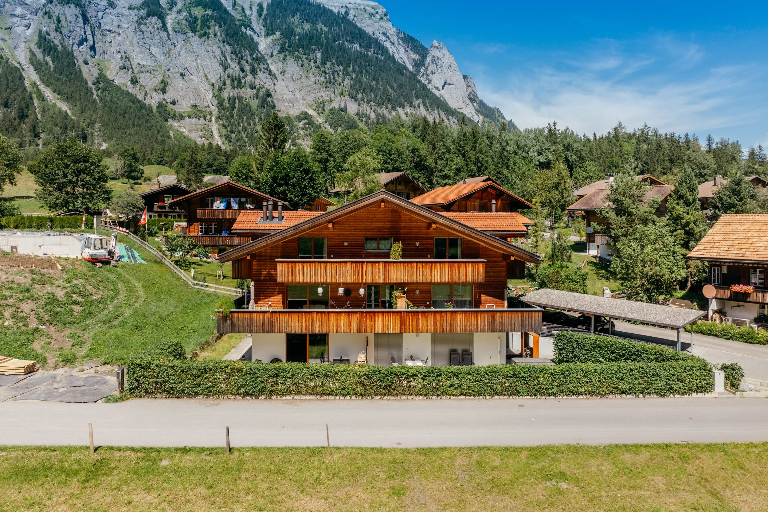 Ein modernes Holzhaus in einer alpinen Umgebung mit Bergen im Hintergrund, umgeben von anderen Holzhäusern, grünem Garten und Bäumen.