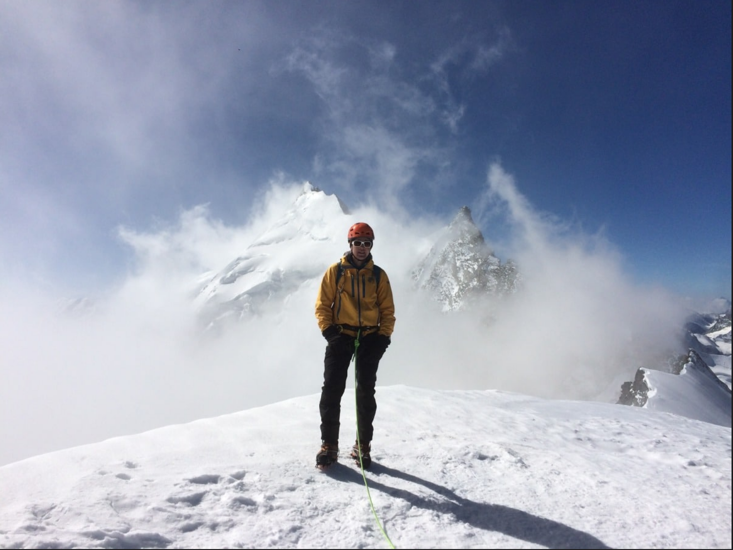Bergsteiger in Schneelandschaft mit hohen schneebedeckten Gipfeln im Hintergrund, trägt gelbe Jacke, Helm und Sonnenbrille, hält Seil in Hand.