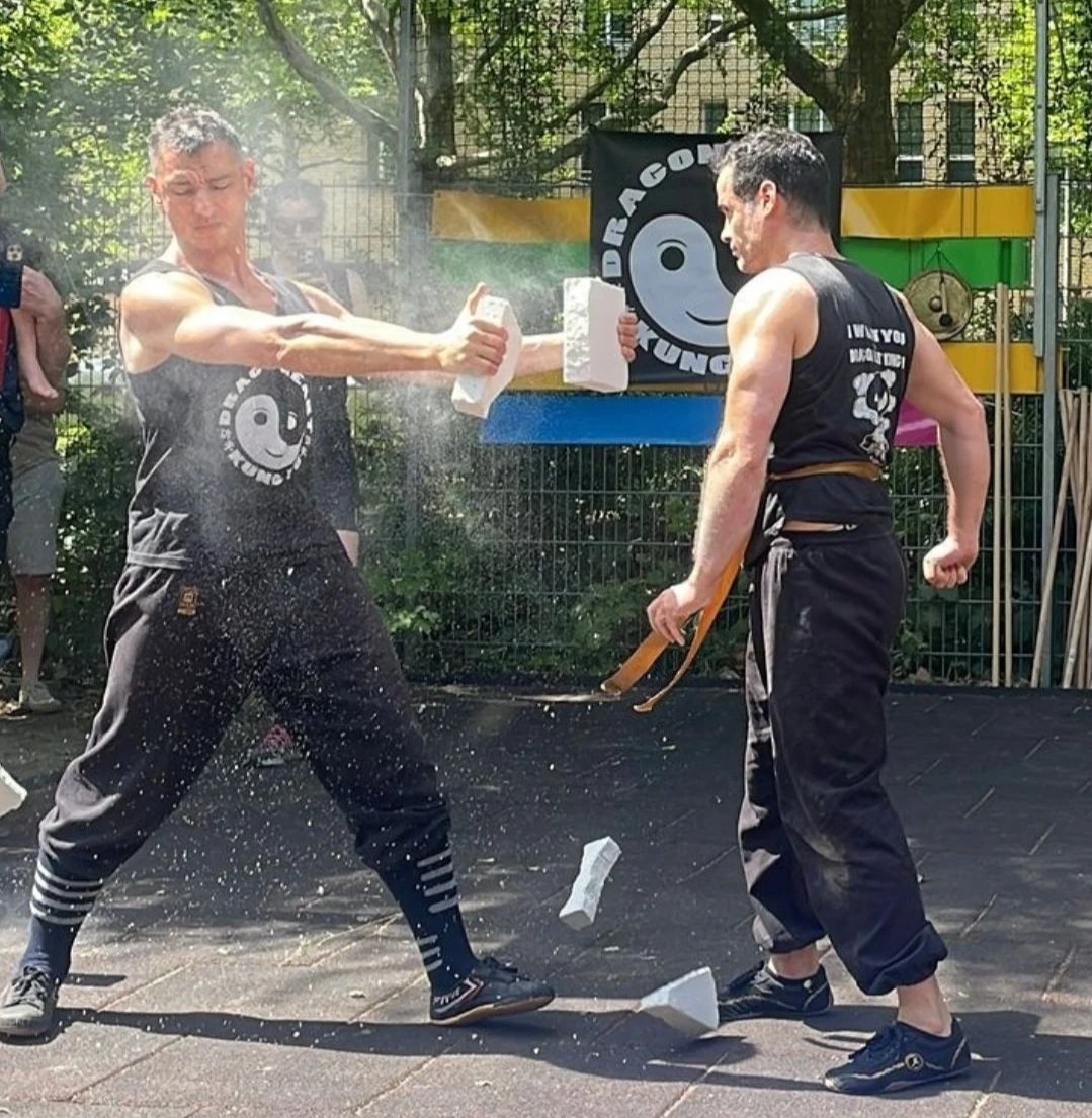Two men practicing martial arts outdoors, with one smashing a concrete block held by the other using a hand strike, resulting in debris and dust.