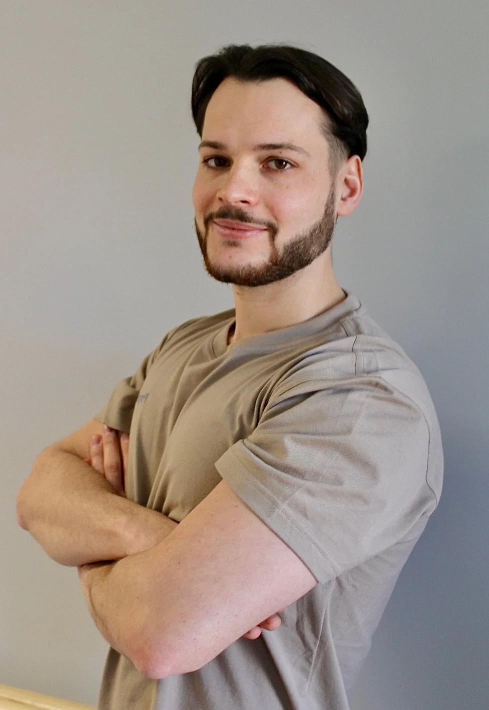 Ein Mann mit dunklem Haar, Bart und mustert, trägt ein graues T-Shirt und steht vor einer grauen Wand.