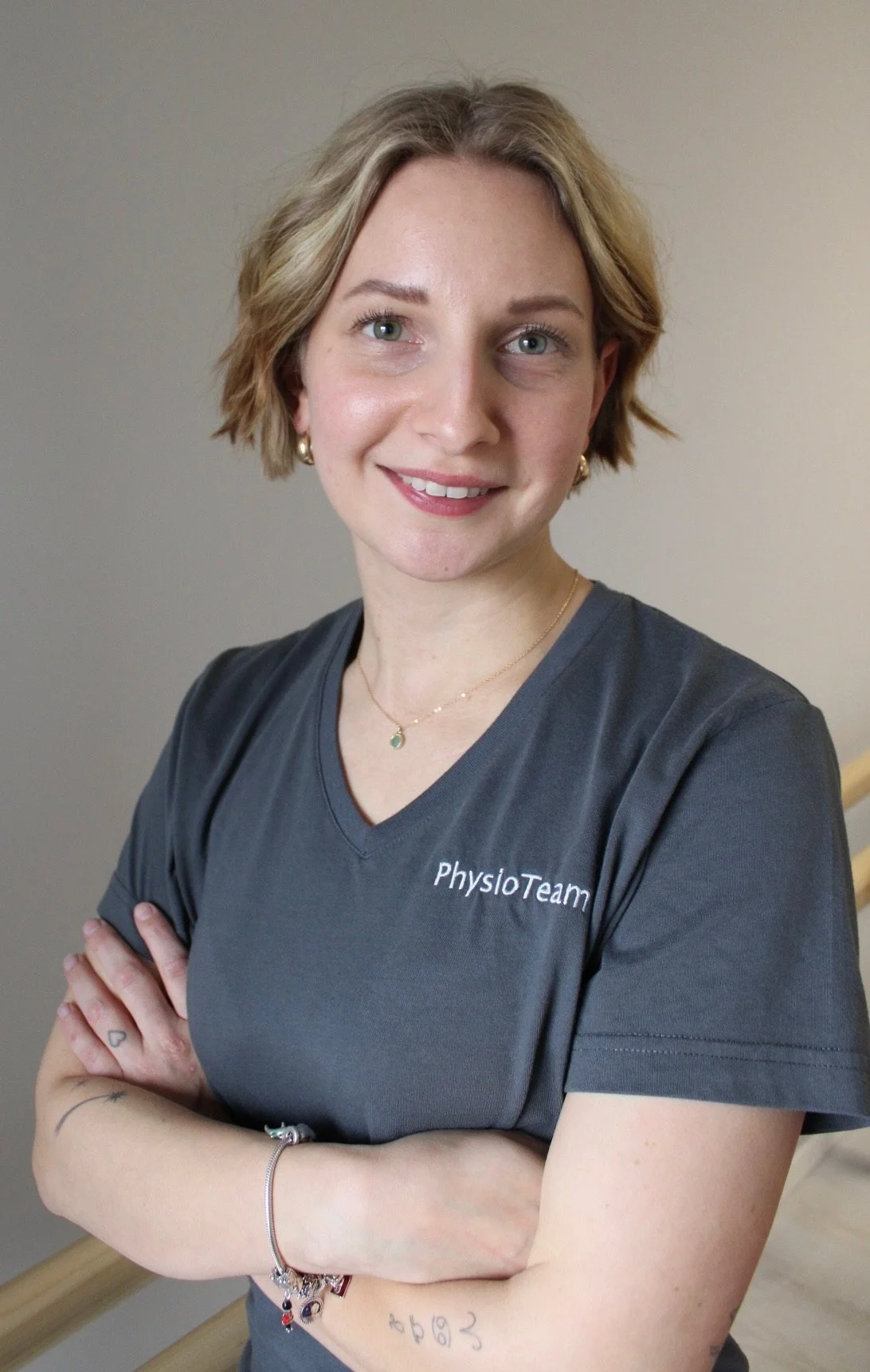 Eine Frau mit kurzen, lockigen blonden Haaren trägt ein graues T-Shirt mit der Aufschrift 'PhysioTeam' und lächelt in die Kamera.