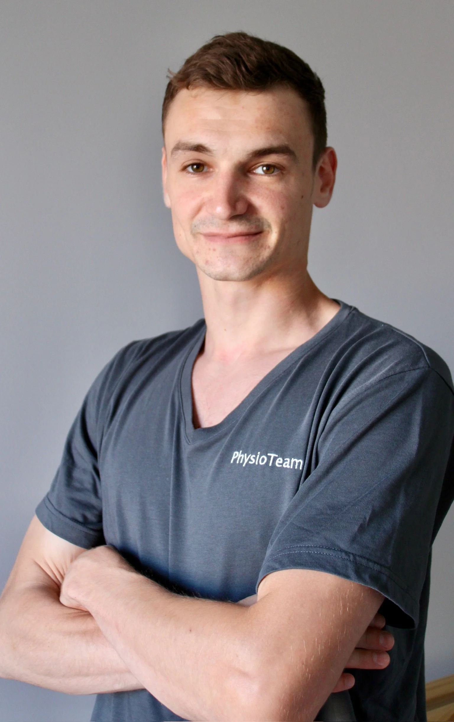 Mann trägt ein graues T-Shirt mit der Aufschrift 'PhysioTeam' und steht vor einem grauen Hintergrund.