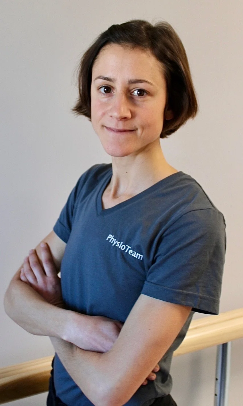 Frau mit kurzen braunen Haaren, blaunem T-Shirt, steht mit verschränkten Armen an einem Geländer.