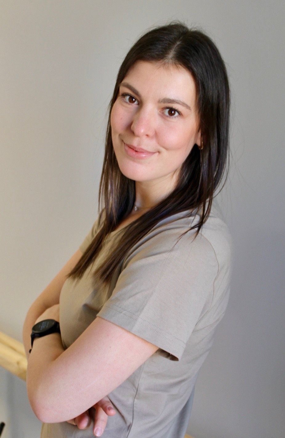 Junge Frau mit dunklen Haaren, lächelnd, trägt eine beige Arbeitsuniform und steht vor einer neutralen Hintergrund. Sie hat die Arme verschränkt und trägt eine Uhr am linken Handgelenk.