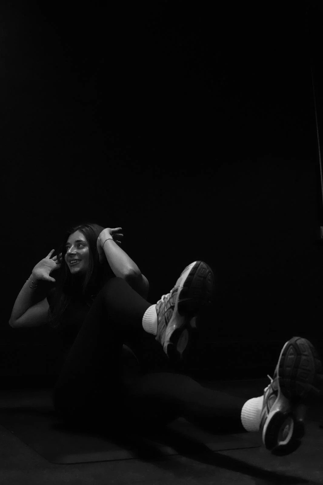 A young woman lying on her back on a dark floor, smiling, with her legs raised and crossed at the ankles, wearing sneakers and dark clothing. She has her hands near her head, in a playful pose, in a dimly lit room.