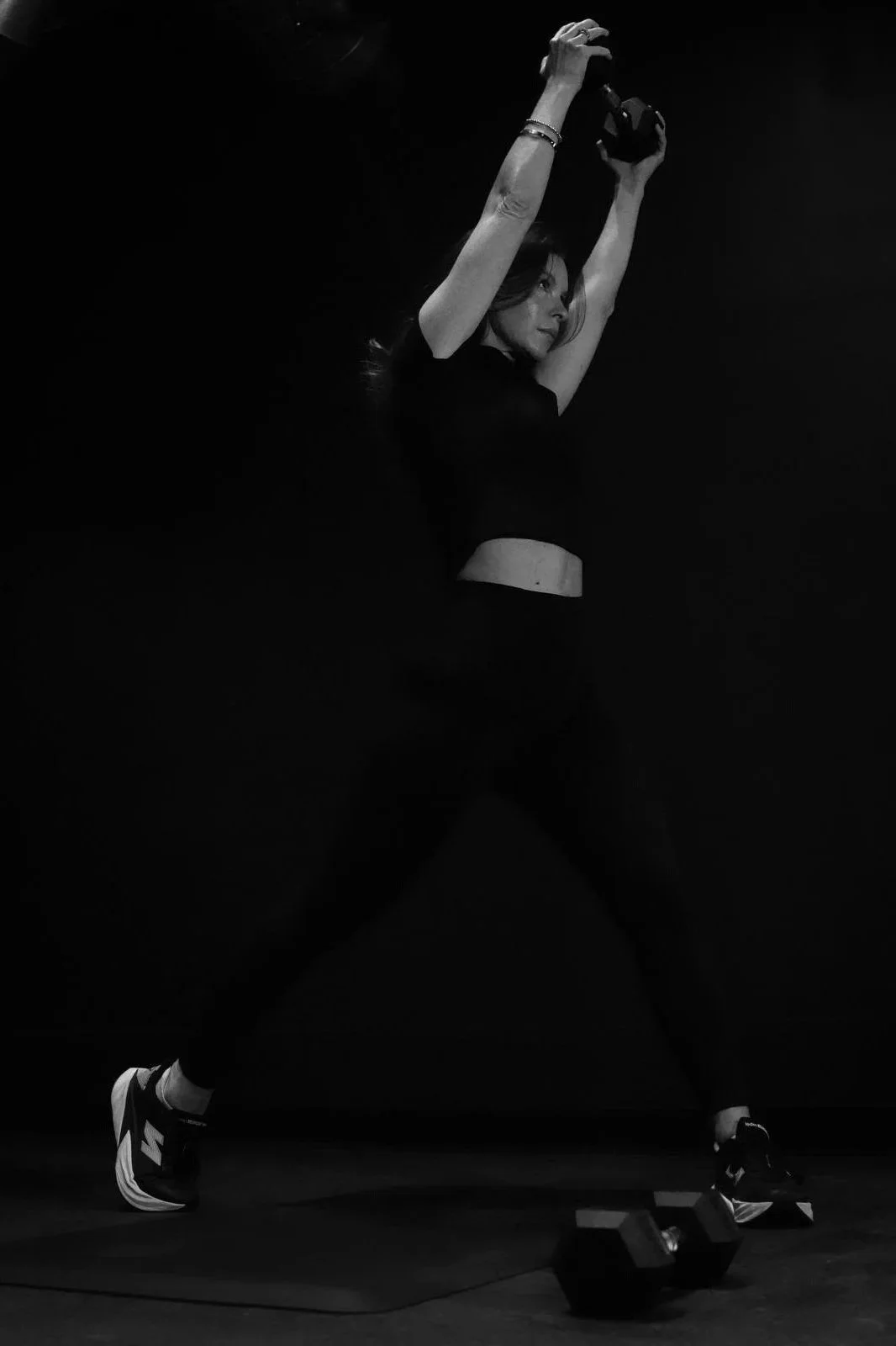 A woman in athletic wear holding a weight above her head during a workout in a dark room, with weights on the floor nearby.
