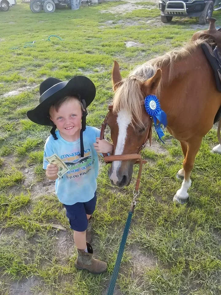 A young boy wearing a large black cowboy hat, blue t-shirt, and cowboy boots holds a 20-dollar bill and stands next to a brown and white horse with a blue ribbon on its bridle. The boy is smiling and the horse is grazing on the grass.