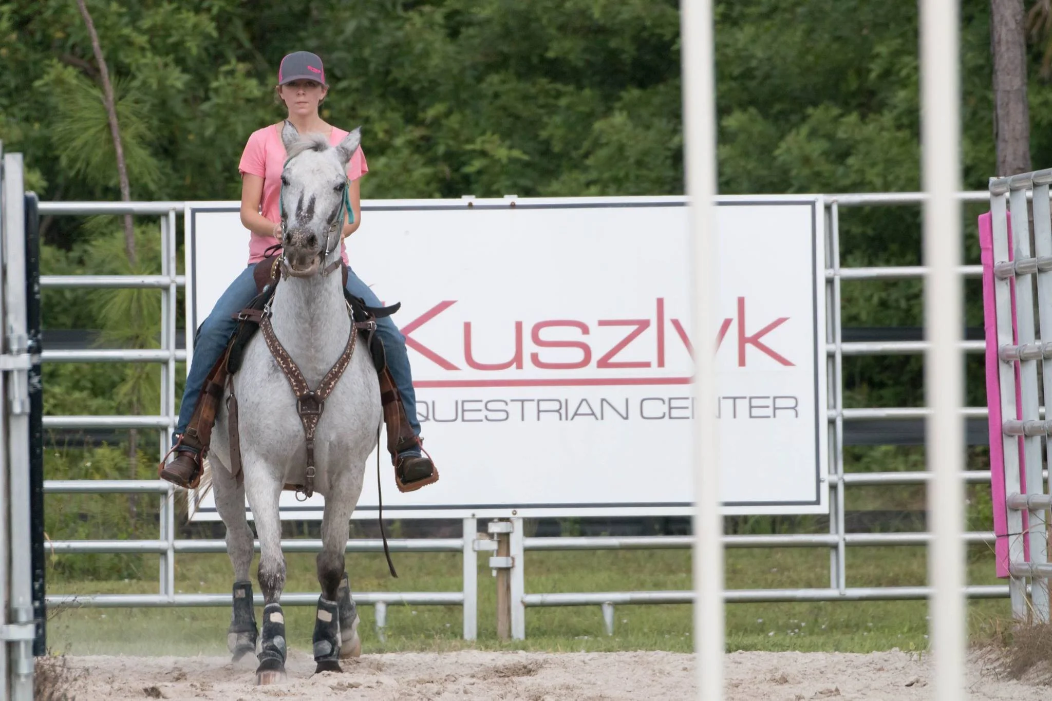 A young girl riding a white horse with black spots on its legs at a horseback riding arena with a sign reading "Kuszyk Equestrian Center" in the background.